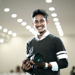 pexels-photo-20166448-20166448 Confident young man holding camera against a backdrop of modern lights, smiling warmly.